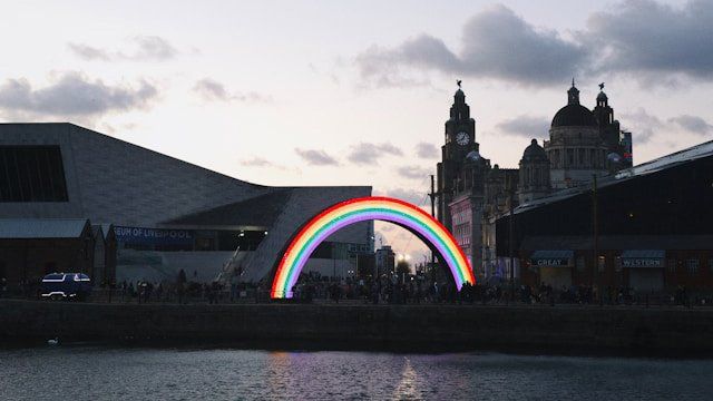 A glowing rainbow arch at dusk beside the Museum of Liverpool and the Royal Liver Building.